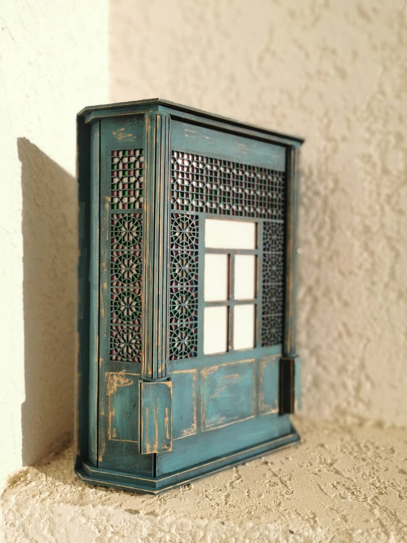 K&ouml;nnte beinhalten: Ein blaugr&uuml;ner Holzschrank mit einem dekorativen Gitterfenster und einer abgenutzten Oberfl&auml;che. Der Schrank hat ein kleines Regal und eine get&auml;felte Unterseite.