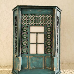 K&ouml;nnte beinhalten: Ein blaugr&uuml;ner Holzschrank mit einem dekorativen Buntglasfenster und einer abgenutzten Oberfl&auml;che. Der Schrank hat ein geometrisches Muster und einen wei&szlig;en Hintergrund.