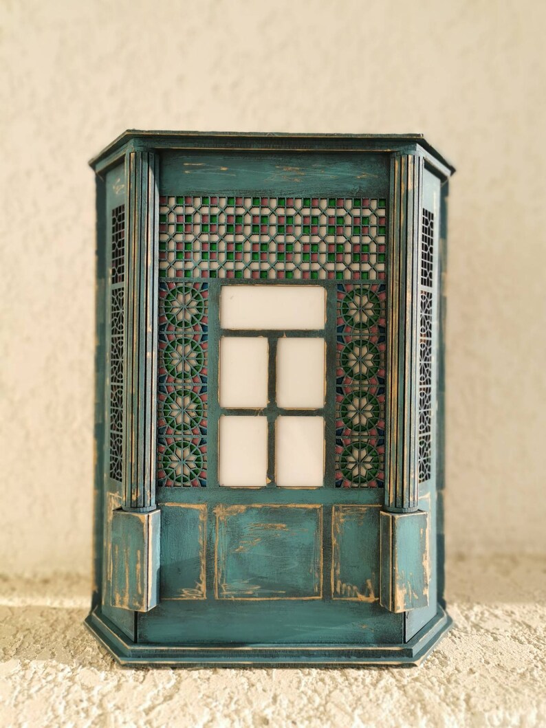K&ouml;nnte beinhalten: Ein blaugr&uuml;ner Holzschrank mit einem dekorativen Buntglasfenster und einer abgenutzten Oberfl&auml;che. Der Schrank hat ein geometrisches Muster und einen wei&szlig;en Hintergrund.