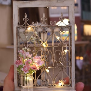 Puede incluir: Una ventana en miniatura de color blanco con un diseño de puerta de metal y un pequeño jarrón con flores rosas. El marco de la ventana tiene la palabra "LAMPINO" impresa en la parte inferior.