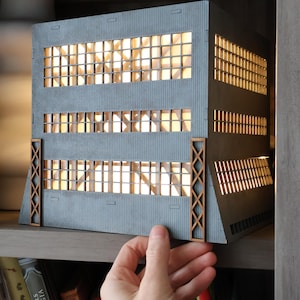 May include: A grey architectural model of a building with multiple rows of windows. The model has a detailed facade with wooden accents and is illuminated from within, creating a warm glow. The model is displayed on a bookshelf.