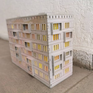 May include: A white model of a multi-story apartment building with windows that have a warm yellow glow. The building is made of cardboard and has a simple design.