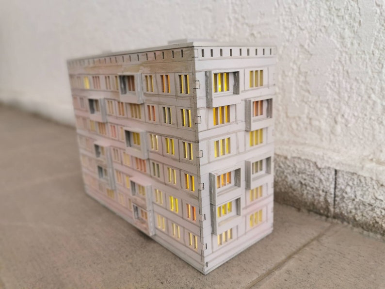 May include: A white model of a multi-story apartment building with windows that have a warm yellow glow. The building is made of cardboard and has a simple design.