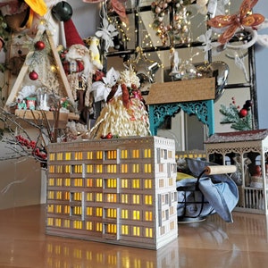 May include: A white cardboard model of a multi-story building with windows that light up. The building is on a wooden table with other decorative items.