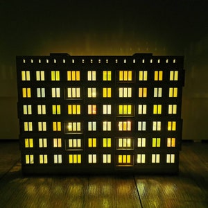 May include: A model of a multi-story apartment building with windows lit up in various shades of yellow and orange. The building is made of dark wood and has a simple design.
