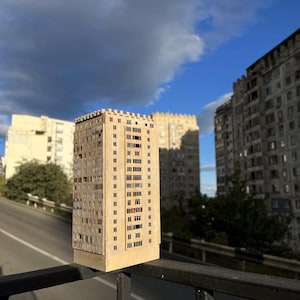 Può includere: Modello in miniatura di un edificio a più piani beige con numerose finestre e un tetto piatto. Le finestre hanno cornici scure. Lo sfondo mostra altri edifici e un cielo nuvoloso.