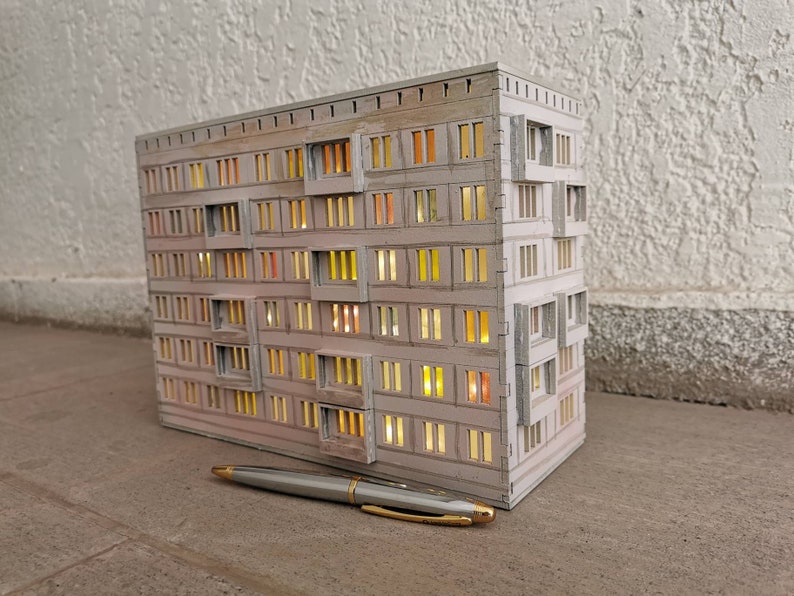 May include: A white cardboard model of a multi-story apartment building with windows that have glowing yellow lights inside. The model is sitting on a light brown tiled surface with a silver pen lying next to it.