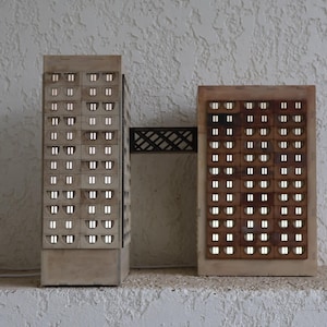 Op de afbeelding: Twee verlichte lampen in architecturale stijl. Eén is een hoge, rechthoekige structuur die lijkt op een gebouw met meerdere verlichte ramen. De andere is een kortere, rechthoekige lamp met een raster van verlichte openingen.