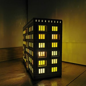 May include: A wooden model of a building with lit windows. The building is dark brown and has many windows with yellow lights. The model is sitting on a wooden floor.