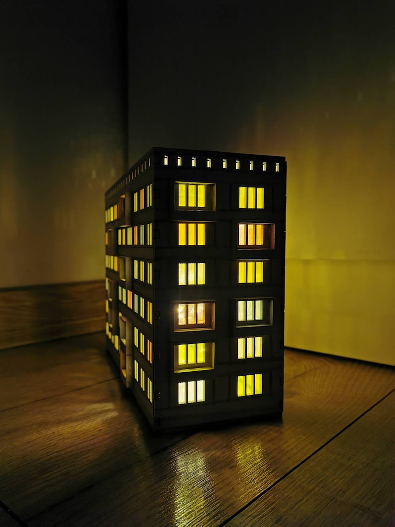 May include: A wooden model of a building with lit windows. The building is dark brown and has many windows with yellow lights. The model is sitting on a wooden floor.