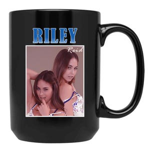 May include: Black ceramic mug featuring the name "RILEY" in blue text above a photo of a woman. The photo is in a white frame and includes the name "Reid". The mug has a large handle.