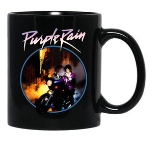 May include: A black ceramic mug featuring the text Purple Rain in a stylized font above a circular graphic. The image depicts a person in a purple jacket sitting on a motorcycle in front of a building with warm yellow light and smoke effects.