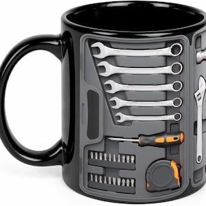 May include: A black ceramic mug featuring a graphic print of a tool organizer case. The design displays silver wrenches, a screwdriver with an orange and black handle, a set of small drill bits, and a tape measure arranged in a gray molded tray.
