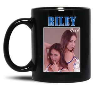 May include: Black ceramic mug featuring the name "RILEY" in blue text. The mug displays a photo of two women, with the name "Reid" printed above the image. The mug has a curved handle.