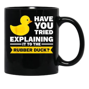 May include: Black ceramic mug with a yellow rubber duck graphic and the text "Have You Tried Explaining It To The Rubber Duck?" The mug has a curved handle and is suitable for hot or cold beverages.