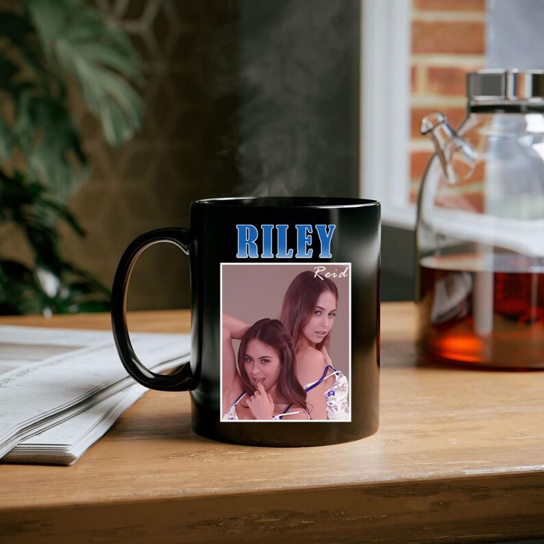 May include: Black ceramic mug featuring the name "RILEY" in blue lettering above a photo of a woman. The photo is in a white frame and the name "Reid" is written in cursive. The mug is on a wooden surface.