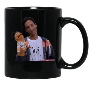 May include: A black ceramic mug featuring a printed image of a person wearing a white t-shirt with a black panda graphic and a multi-colored zip-up hoodie, holding a tan hand puppet wearing a striped shirt on their right hand.