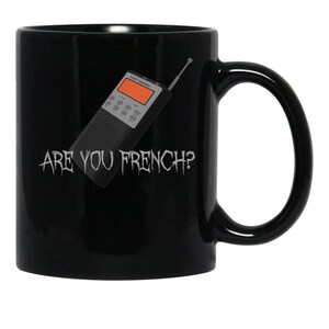 May include: A black ceramic mug featuring a graphic of a grey spirit box device with an orange screen and an antenna. Below the device, the text ARE YOU FRENCH? is printed in a distressed, jagged white font against the solid black background.