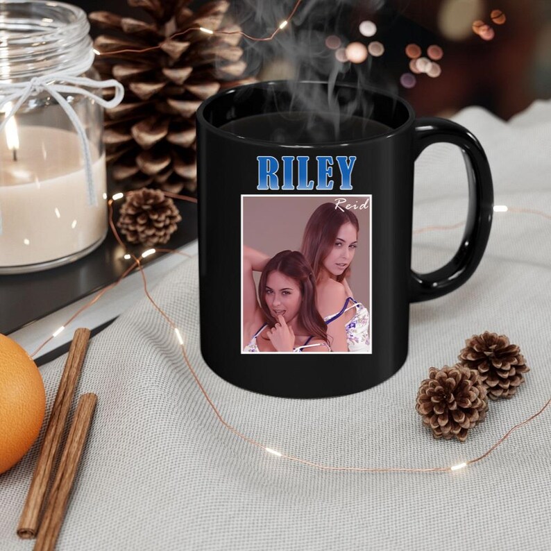May include: Black ceramic mug with a large handle. The mug features the name "RILEY" in blue text above a photo of two women. The name "Reid" is below the photo. Steam rises from the mug, suggesting a hot beverage.