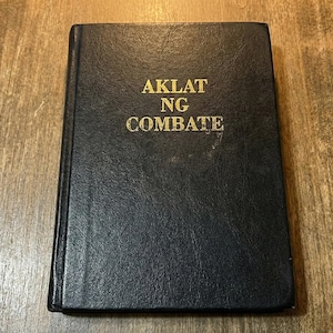 May include: A black book with the words "AKLAT NG COMBATE" in gold lettering. The book is closed and resting on a wooden surface. The cover has a textured appearance.