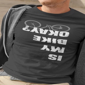 以下が含まれることがあります： 白い文字で「Is My Bike Okay?」と書かれた濃いグレーのTシャツ。テキストの上に自転車のグラフィックがあります。