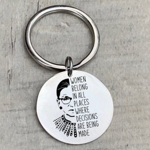 Op de afbeelding: Zilveren sleutelhanger met een rond label met een zwart-wit afbeelding van Ruth Bader Ginsburg en de tekst "Vrouwen horen op alle plekken waar beslissingen worden genomen."