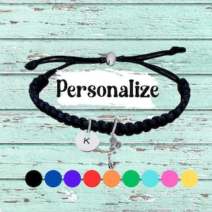 May include: Black braided bracelet with a silver charm and a round pendant with the letter "K". The bracelet has a sliding knot closure. The word "Personalize" is written in black letters on the image. A row of colorful circles is at the bottom.