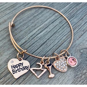 May include: A gold bangle bracelet with charms. The charms include a heart with the text "Happy Birthday", a number 2, a wine glass, a heart with rhinestones, and a pink rhinestone.
