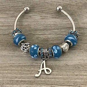 May include: A silver charm bracelet with blue and white beads and a silver charm in the shape of the letter A.