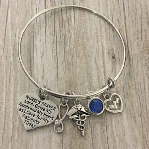 May include: Silver-toned bangle bracelet with multiple charms. Charms include a heart with the text "NURSE'S PRAYER", a stethoscope, a medical symbol, a blue crystal, and a heart with a lightning bolt. The bracelet is on a wooden surface.