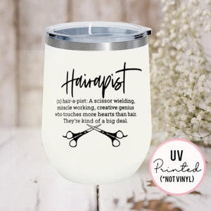 May include: White stainless steel wine tumbler with a black design that says "Hairapist (n) hair-a-pist: A scissor wielding, miracle working, creative genius who touches more hearts than hair. They're kind of a big deal." with a pair of scissors crossed underneath the text. The tumbler has a black lid and a pink sticker on the side that says "UV Printed (*NOT VINYL)"