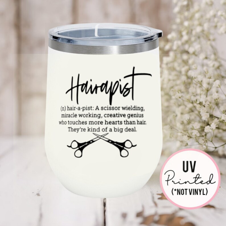 May include: White stainless steel wine tumbler with a black design that says "Hairapist (n) hair-a-pist: A scissor wielding, miracle working, creative genius who touches more hearts than hair. They're kind of a big deal." with a pair of scissors crossed underneath the text. The tumbler has a black lid and a pink sticker on the side that says "UV Printed (*NOT VINYL)"