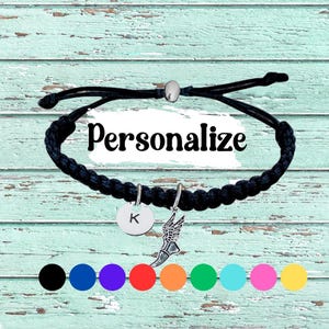 May include: Black braided bracelet with a silver charm featuring a running shoe and a round charm with the letter "K". The word "Personalise" is displayed on the bracelet. A selection of colour options are shown below the bracelet.