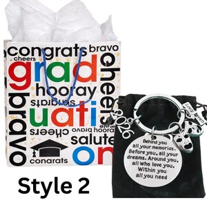 May include: A graduation gift set featuring a gift bag with graduation-themed text and a keychain. The bag has a blue handle and is filled with white tissue paper. The keychain has a round charm with an inspirational message.