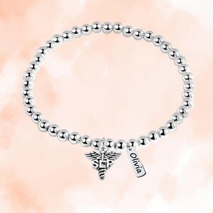 May include: Silver beaded bracelet with a charm of a medical caduceus with the letters "SLP" and a small tag that says "Olivia".