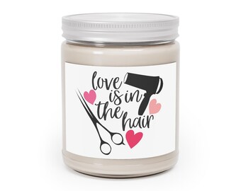 May include: A beige-colored candle in a glass jar with a silver lid. The label features the phrase "love is in the hair" with a black hairdryer, scissors, and pink hearts. A decorative item for a salon or hairstylist.