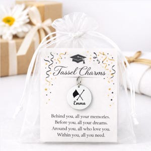 May include: A white graduation tassel charm with the name "Emma" and a crossed flag design. The charm hangs from a card with the words "Tassel Charms" and a graduation cap graphic. The charm is packaged in a white organza bag.