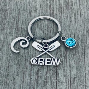 May include: Silver-toned keychain with a nautical theme. The keychain features a large ring with three charms: a wave, crossed oars, and a blue crystal. The word "CREW" is displayed below the oars.
