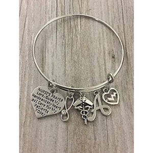 May include: Silver charm bracelet with a heart charm that says "Nurse's Prayer, Lord, Guide My Hands and My Heart as I Care for My Patients Today". The bracelet also has a stethoscope charm, a caduceus charm, and a letter A charm.