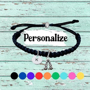 May include: Black braided bracelet with a silver charm of ballet shoes and a round charm with the letter "K". The word "Personalize" is written across the image. A selection of color options are shown below the bracelet.