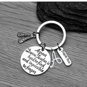 May include: Silver keychain with a charm that reads "A great dance teacher is hard to find and impossible to forget". The keychain also has a charm with the letter "A" and a charm that reads "Thank you".