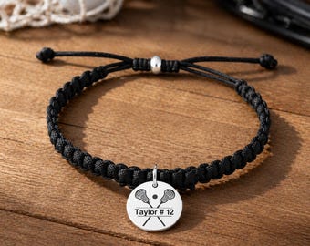 May include: Black braided bracelet with a silver charm. The charm features crossed lacrosse sticks, a lacrosse ball, and the text "Taylor #12". The bracelet is on a wooden surface.