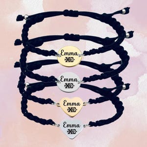 May include: Four black cord bracelets with silver and gold charms. Each charm has the name "Emma" and a cross with an arrow engraved on it. Two charms are round and two are heart shaped.