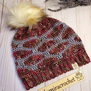 May include: A crocheted beanie with a multi-colored pattern in shades of red, brown, and gray. The beanie has a fluffy pompom on top and a wooden tag attached to the side that says "@lumiracrochet".