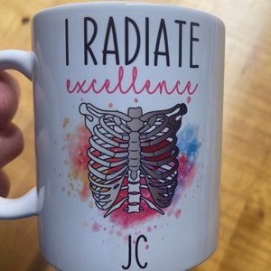 Könnte beinhalten: Eine weiße Keramiktasse mit dem Text "I radiate excellence" und einer bunten Illustration eines Brustkorbs mit einem Herzen. Die Tasse ist mit den Initialen "JC" personalisiert.
