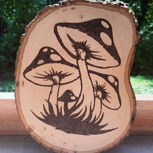 Puede incluir: Una rodaja de madera con borde de corteza natural presenta un diseño pirograbado de tres setas con sombreros punteados y tallos detallados. Las setas se colocan sobre un fondo de hierba estilizada, todo quemado en la madera clara.