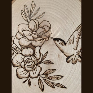 May include: Wood-burned art piece featuring a hummingbird and flowers. The hummingbird is depicted with detailed wings and a long beak, reaching towards the flowers. The flowers and leaves are intricately designed with a dark brown burn, set against a light wood background.