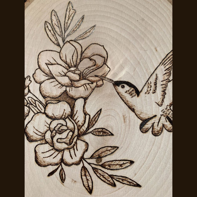 May include: Wood-burned art piece featuring a hummingbird and flowers. The hummingbird is depicted with detailed wings and a long beak, reaching towards the flowers. The flowers and leaves are intricately designed with a dark brown burn, set against a light wood background.