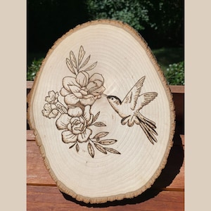 May include: A wood slice with a hummingbird and floral design, created using pyrography. The hummingbird is depicted in flight, reaching towards the flowers. The wood has a natural, light brown color with visible grain and bark.