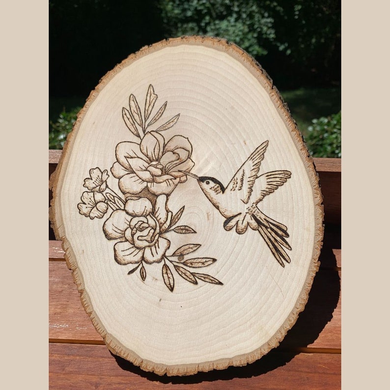May include: A wood slice with a hummingbird and floral design, created using pyrography. The hummingbird is depicted in flight, reaching towards the flowers. The wood has a natural, light brown color with visible grain and bark.